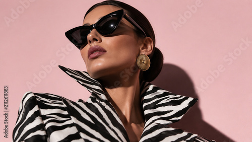 Stylish fashion portrait of a woman wearing a zebra-striped structured jacket, black sunglasses and gold earrings against a soft pink background. Ideal for fashion magazines, eyewear campaigns, luxury
