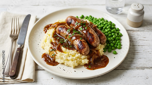 Traditional bangers and mash with onion gravy and green peas on a plate with cutlery and a salt shaker on a white rustic wooden table