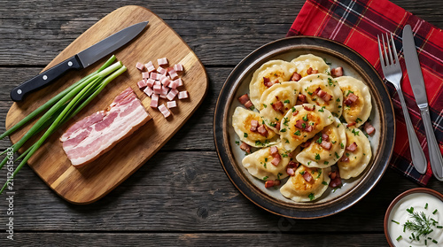 Traditional pierogi dumplings with bacon bits on a plate next to a wooden cutting board with raw bacon, green onions, and a kitchen knife