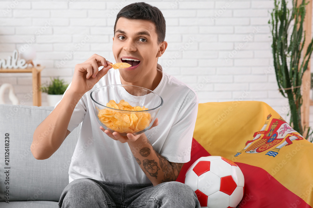 Fototapeta premium Young man with bowl of potato chips, soccer ball and flag of Spain watching football on sofa at home