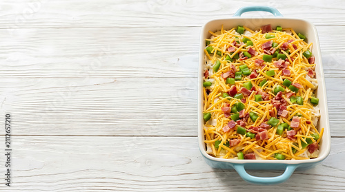 Unbaked casserole dish filled with ingredients topped with shredded cheddar cheese, diced green peppers, and bacon bits on a white wooden table