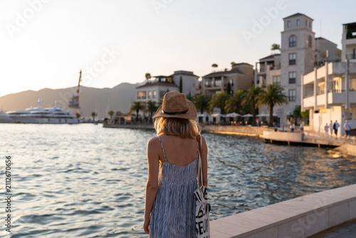 Young woman in straw hat walking along the waterfront in Montenegro at golden hour, with marina, palm trees and coastal architecture in warm evening light
