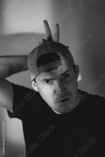 Young man in baseball cap making bunny ears gesture over head. Portrait of playful guy acting silly and looking into camera. Concept of humor, fun lifestyle and casual expression.