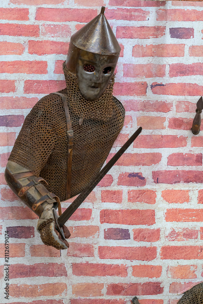 Obraz premium Medieval knight in chainmail stands against brick wall. Helmet with pointed top covers his face. He grips a sword with both hands. Brick wall behind him is textured and red