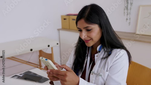 Young Indian female doctor sitting at her desk in a modern clinic office while using her smartphone for telehealth consultation, communication or browsing medical apps