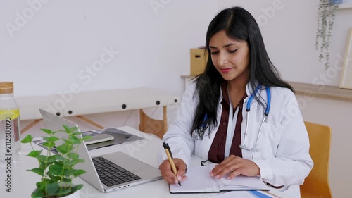 Professional female general practitioner with a stethoscope writing a patient medical prescription in a notebook while sitting at her desk in a modern hospital office or clinic
