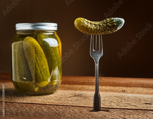 a single green pickle on a silver fork with a glass jar of pickled cucumbers on a rustic wooden table in a home kitchen