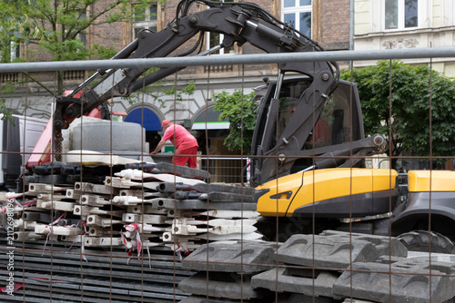 Картината върху платно A construction site behind a metal mesh fence, a yellow and black excavator