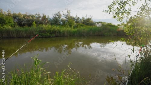 Fishing rods set up along calm riverbank surrounded by green vegetation. Concept of recreational fishing and peaceful outdoor relaxation