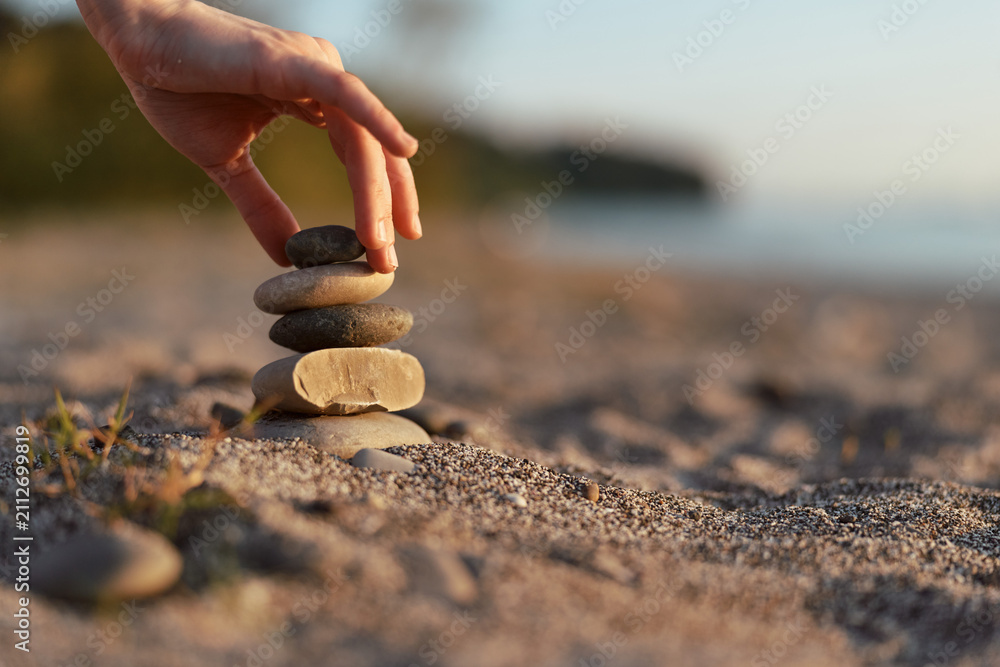 Fototapeta premium Woman lifestyle concept detail where a hand balances smooth zen stones on a sandy beach. Peaceful outdoor meditation scene at sunset light creating a calm and mindful atmosphere.