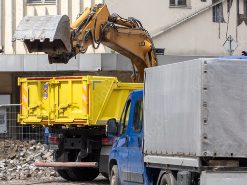 A yellow hydraulic excavator next to a large, yellow RORO (Roll-On/Roll-Off) skip container. commercial waste removal services. A blue truck  for hauling away the debris. Demolition operations. 