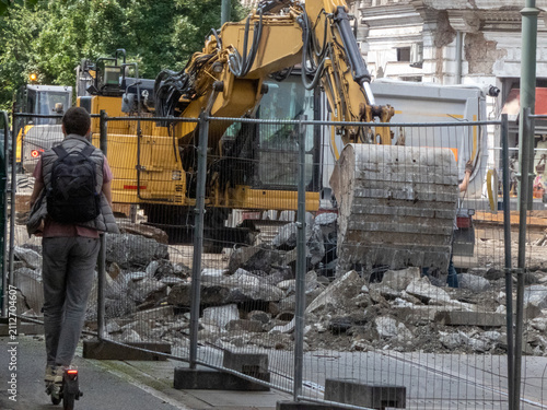 Excavator with a yellow arm. A truck with reflective panels for hauling debris, materials. The area is secured by metal fencing, separating the construction from the surrounding urban environment.