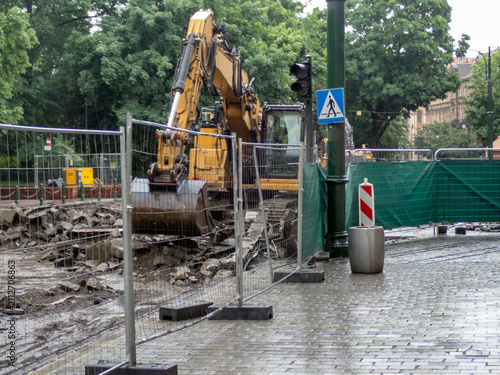 A yellow excavator performing demolition, excavation work behind a temporary metal fence. Rubble, cleared debris are visible. A green mesh screen is attached to the fence to provide safety.