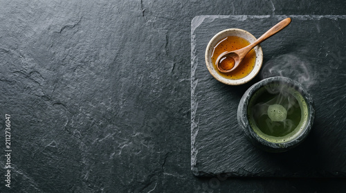 Top view of hot tea and honey in bowls on dark slate board with steam rising against textured black background for food photography.