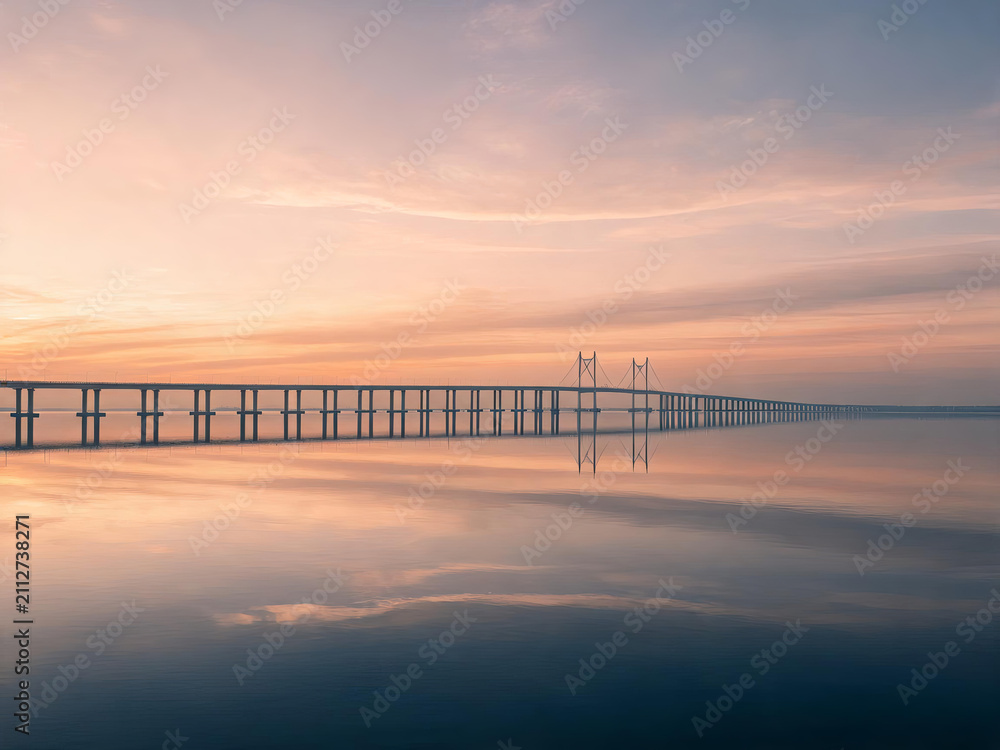 Fototapeta premium A long bridge spans calm water, with clear reflections under a soft sunrise sky, creating a serene landscape.