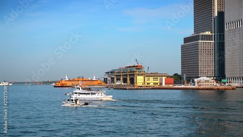 Boats passing South Ferry waterfront in Lower Manhattan. A white tour vessel and small patrol craft leave wakes beside the orange Staten Island Ferry.