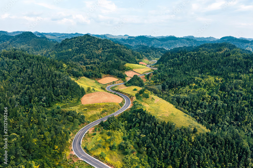Fototapeta premium A winding paved road cuts through dense forests and farmlands in a mountainous natural landscape under a clear sky.