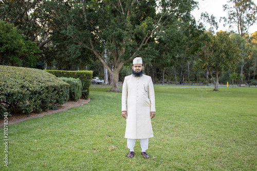 Full-length portrait in traditional formal clothing captured in a spacious green park with tall trees and landscaped surroundings. Natural lighting and open outdoor scenery.