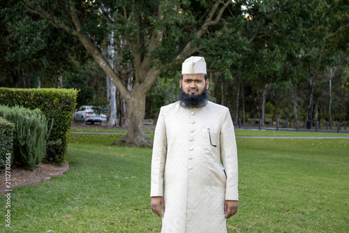 Outdoor portrait of a person wearing elegant cream-colored traditional formal attire, standing beside a tranquil pond in a beautifully landscaped public park