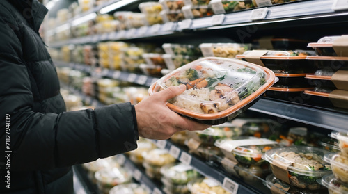 A hand picking up a fresh prepared meal in a plastic container from a refrigerated display case at a grocery store with supermarket shopping customer selection