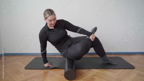 Woman in black sportswear foam rolls her glute and hip on a mat to loosen tight muscles and ease pain after exercise.