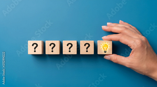 Hand placing wooden block with a glowing lightbulb beside question marks. Problem solving, innovation and idea concept