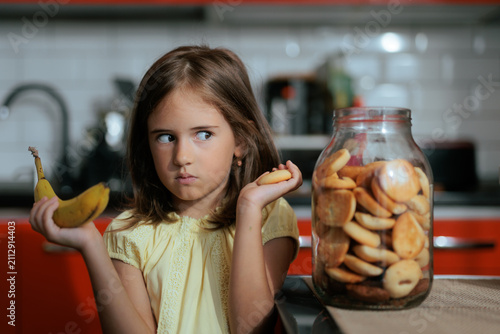 Картината върху платно Little Girl Comparing Cookie And Banana Thinking About Healthy Snack Options