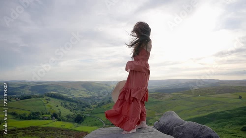 Woman in orange dress stands on hilltop and spins in the wind while enjoying nature in the countryside
