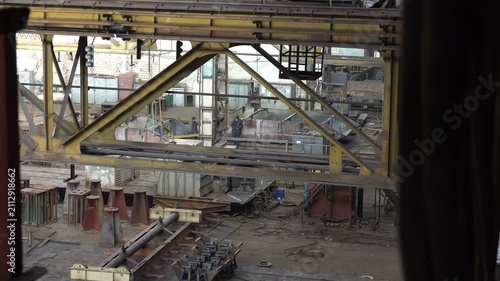 Wide view through a dark doorway into a large industrial workshop with a yellow overhead crane, steel beams and two workers on the dusty floor