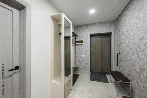 compact apartment entryway features a mirrored storage unit, a dark wood door, a cushioned bench with metal frame, textured grey walls, and light tiled flooring under recessed ceiling lighting