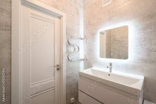 compact bathroom features a white vanity with a rectangular sink, a mirror with LED backlight, a chrome heated rail, and beige‑gray tiled walls