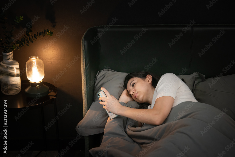 Fototapeta premium Lying in bed woman with insomnia scrolls her phone under warm lamp light creating quiet restless mood in dim bedroom scene