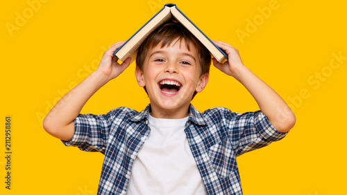 Laughing little boy holding an open book on his head like a roof. Happy child playing against a yellow background. Education and fun learning concept