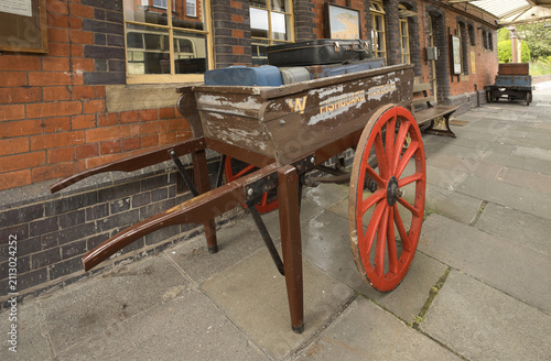Vintage wooden luggage cart with large red spoked wheels holding old suitcases on a brick railway platform.