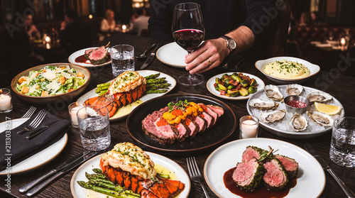 Gourmet Feast: A lavish spread of exquisite dishes, including lobster, prime rib, oysters, and a fresh salad, is meticulously arranged on a dark, elegant table, accompanied by a glass of red wine.