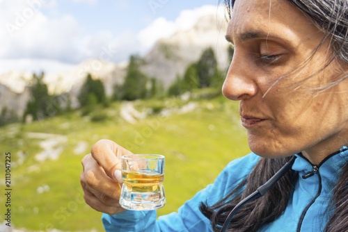 Close up woman traveler drinking local herbal spirit schnapps or grappa shot glass wooden terrace green mountain valley backdrop Dolomites Italy. Fragrant, grape-based pomace home made brandy