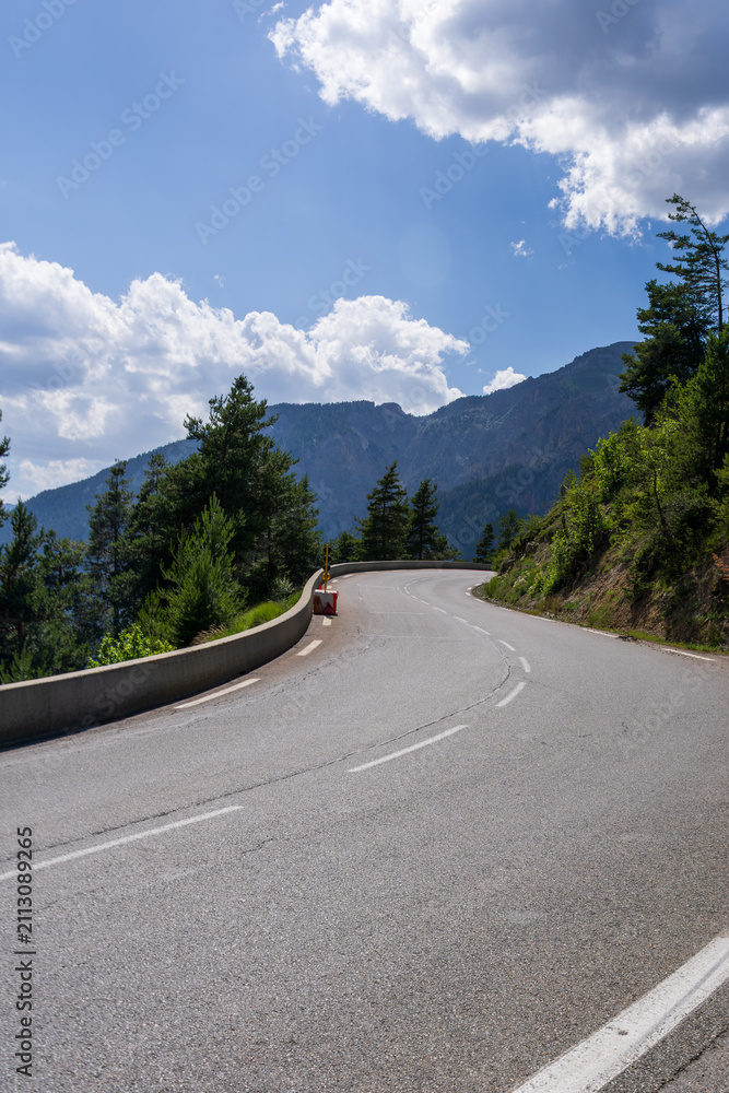 Obraz premium A winding mountain road curves past a concrete barrier and tall pine trees with dramatic peaks rising in the background. Fluffy white clouds fill the vivid blue sky above this scenic alpine drive