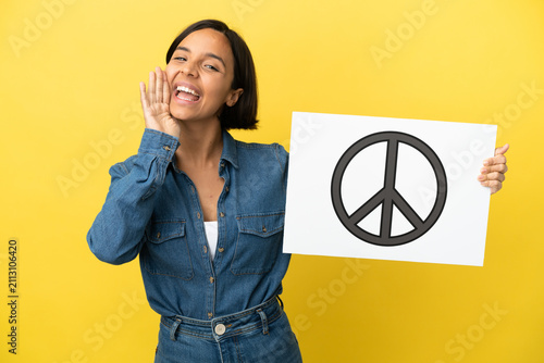 Young mixed race woman isolated on yellow background holding a placard with peace symbol and shouting