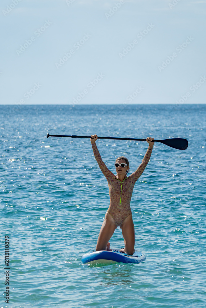 Fototapeta premium Paddleboarding, woman, ocean. Joyful woman kneeling on a paddleboard with paddle raised high, celebrating success during summer vacation, copy space