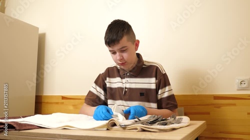 Teenager wearing protective gloves is polishing a metal spoon with a towel while carefully cleaning tableware before table setting.