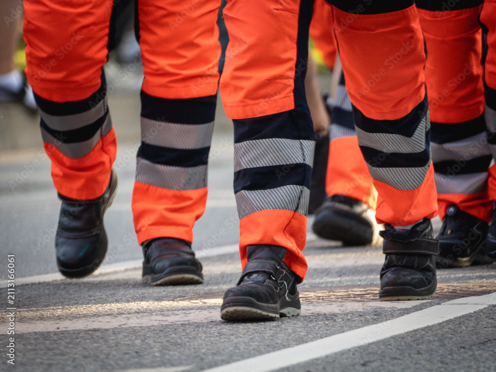 Obraz premium Close-up of emergency service personnel wearing high-visibility orange trousers with reflective bands and black safety boots while marching together