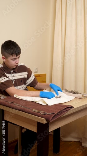 Teenager wearing protective gloves is polishing metal forks with a towel while carefully cleaning tableware before table setting.