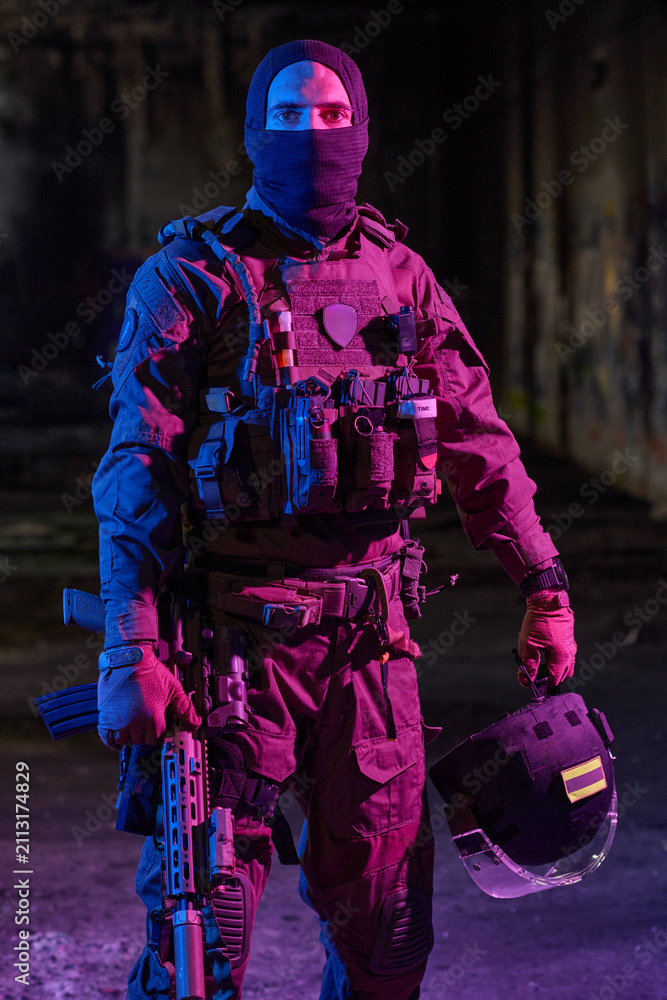 Obraz premium Armed Tactical Soldier In Night Operation Wearing Ballistic Vest Holding Helmet And Rifle