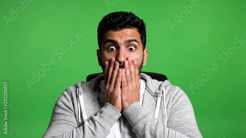 Shocked young man covering his mouth with both hands in absolute disbelief against a green background