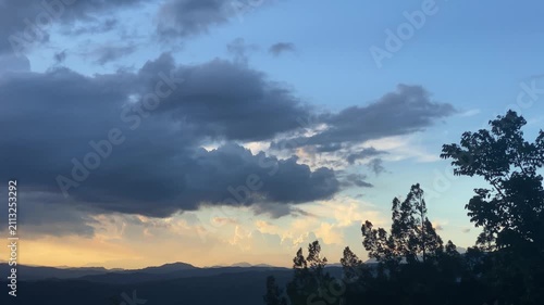 Silhouetted trees against the glowing horizon, where clouds are dispersed across the sky, catching the sunlight in warm orange and yellow hues, leading to a clear blue sky.