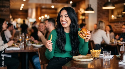 Cheerful young woman eating burger and fries in restaurant, happy female customer enjoying fast food meal, casual dining lifestyle in busy cafe