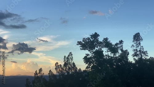 Timelapse of Silhouetted trees against the glowing horizon, where clouds are dispersed across the sky, catching the sunlight in warm orange and yellow hues, leading to a clear blue sky.