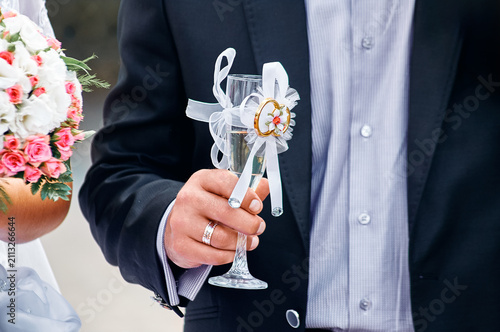 Foto Groom in a suit holds a ribboned champagne glass next to the bride’s pink and white bouquet, capturing a celebratory wedding moment of rings, romance and elegant floral details