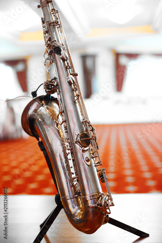 Photography A close-up of a polished silver saxophone on a stand in a brightly lit ballroom, evoking jazz performance, musical practice, elegance and warm reflective tones suitable for concerts and events