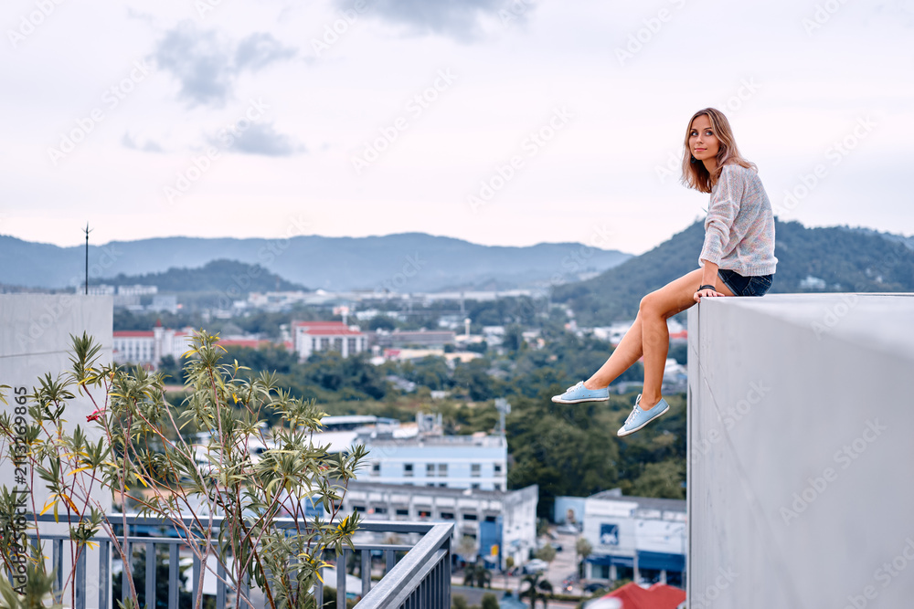 Obraz premium Young woman sits on a rooftop edge with a relaxed, contemplative expression while looking over a cityscape and distant mountains, conveying freedom, adventure, urban lifestyle and quiet reflection.